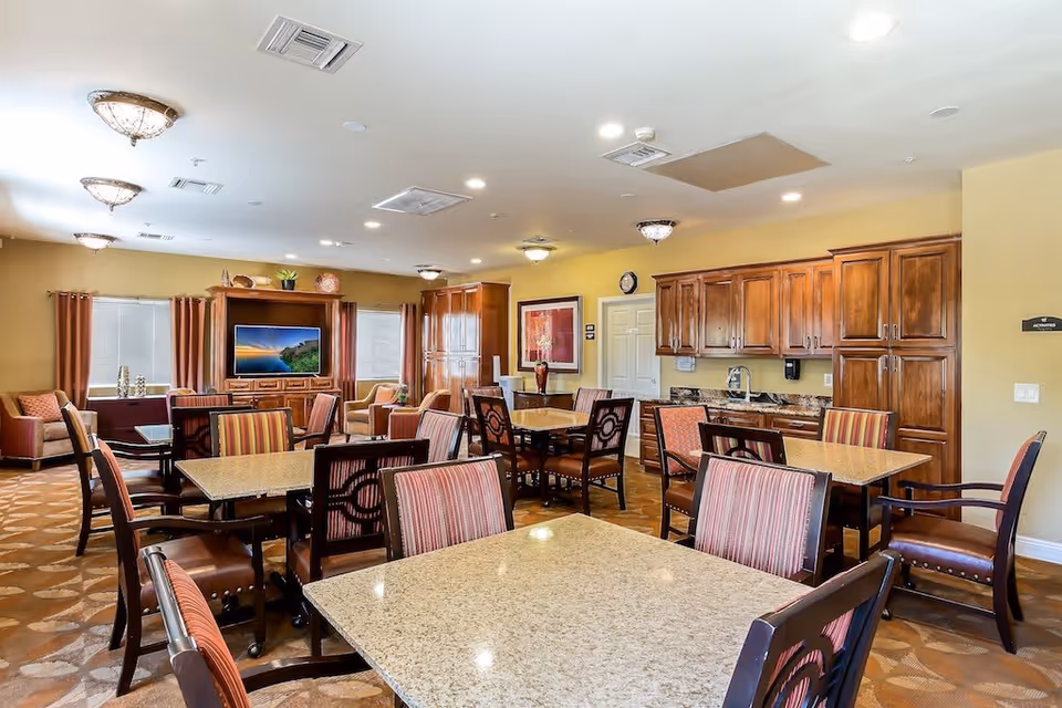 A spacious, well-lit common area with multiple tables and chairs arranged for group seating. The room features warm yellow walls, wooden cabinetry with a countertop and sink, and a large flat-screen TV mounted on a wooden entertainment center. There are windows with curtains, decorative ceiling lights, and framed artwork on the walls.