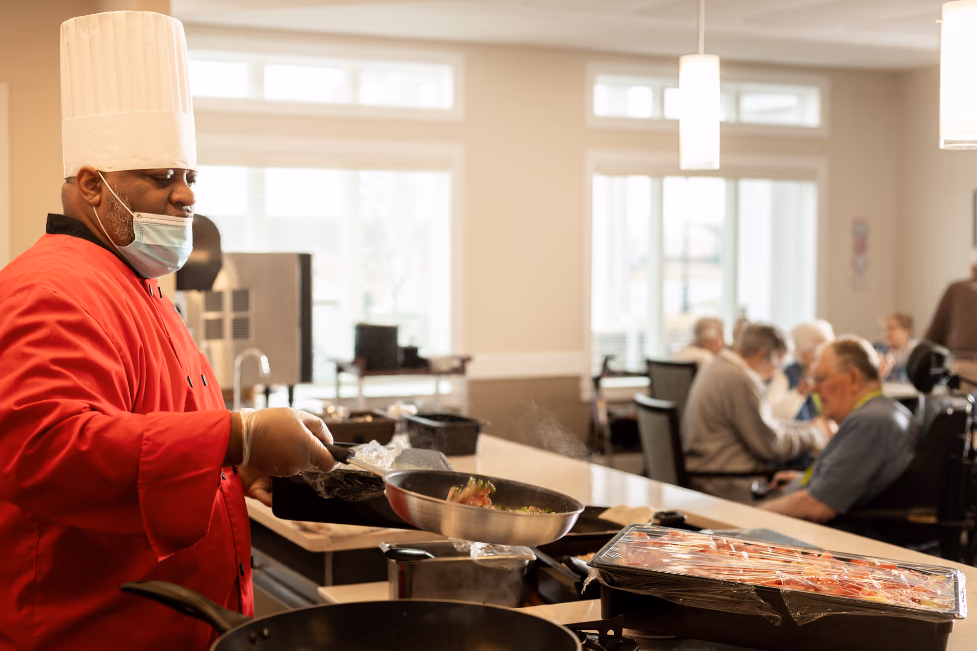 A chef wearing a red uniform, white chef hat, and face mask is cooking food in a pan in a kitchen area. In the background, elderly people are seated and engaged in conversation in a well-lit dining or common area with large windows.