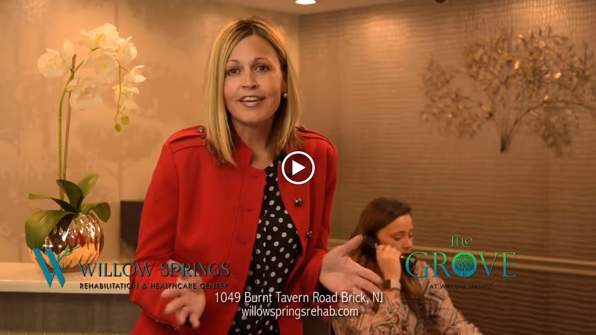 A woman in a red jacket speaking in an indoor setting with a reception desk and a potted orchid plant. Another woman is seated behind the desk, talking on the phone. The background features a textured wall with decorative elements.