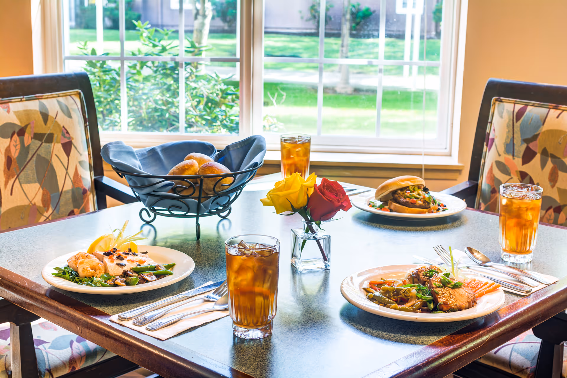 A dining table set with plated meals, glasses of iced tea, a bread basket, and a small vase of roses by a window.