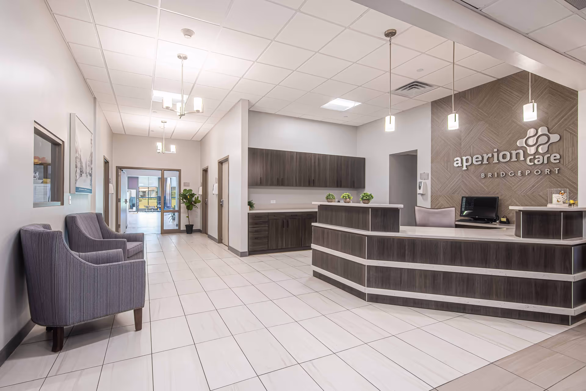 Reception area of a senior living facility with a modern front desk, three pendant lights hanging from the ceiling, and a sign on the wall that reads 'aperion care BRIDGEPORT'. There are two gray armchairs along the left wall, a potted plant near the glass entrance doors, and cabinets behind the reception desk.