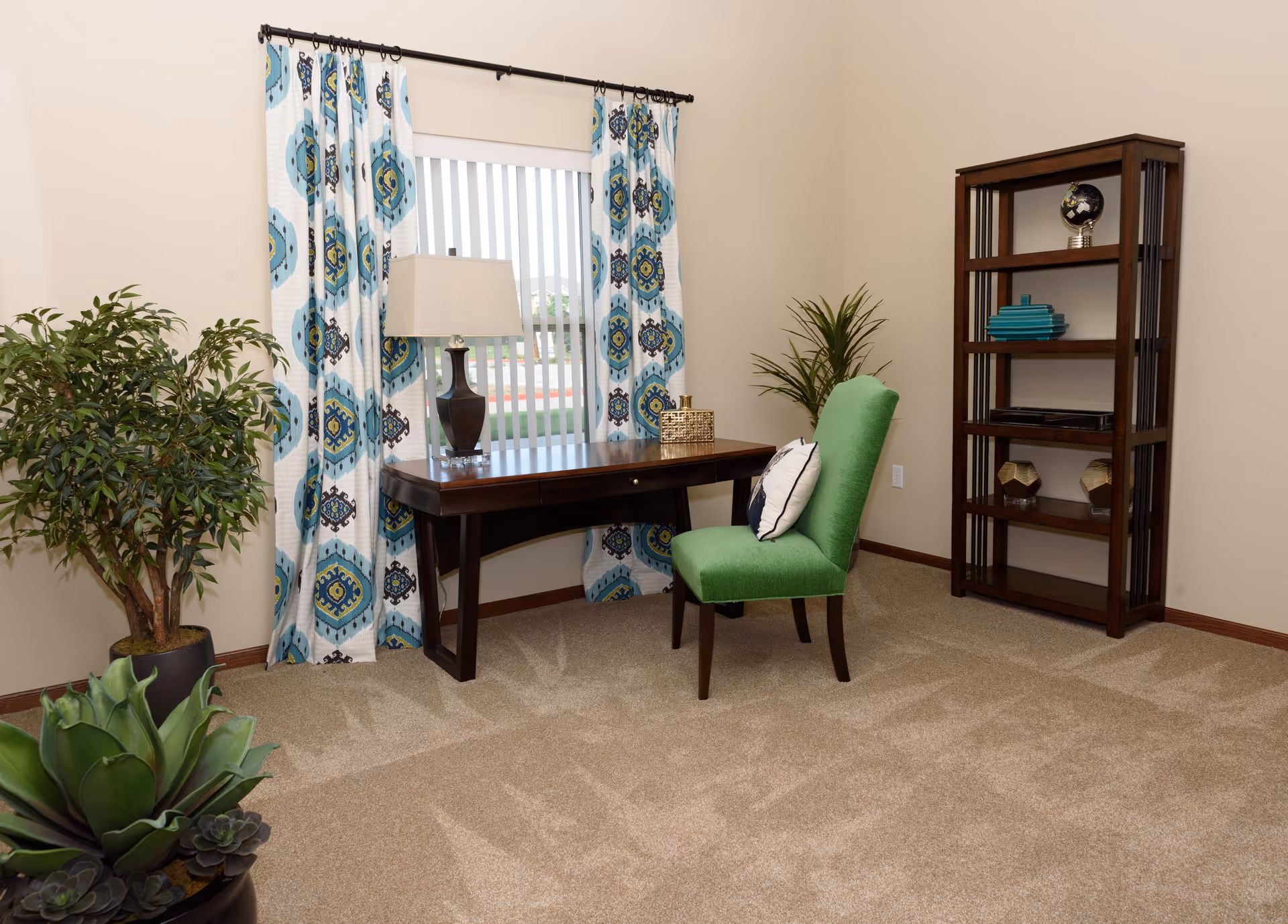 Light-filled sitting room with a dark wood desk, green upholstered chair, patterned curtains, potted plants, and a tall wooden bookshelf.