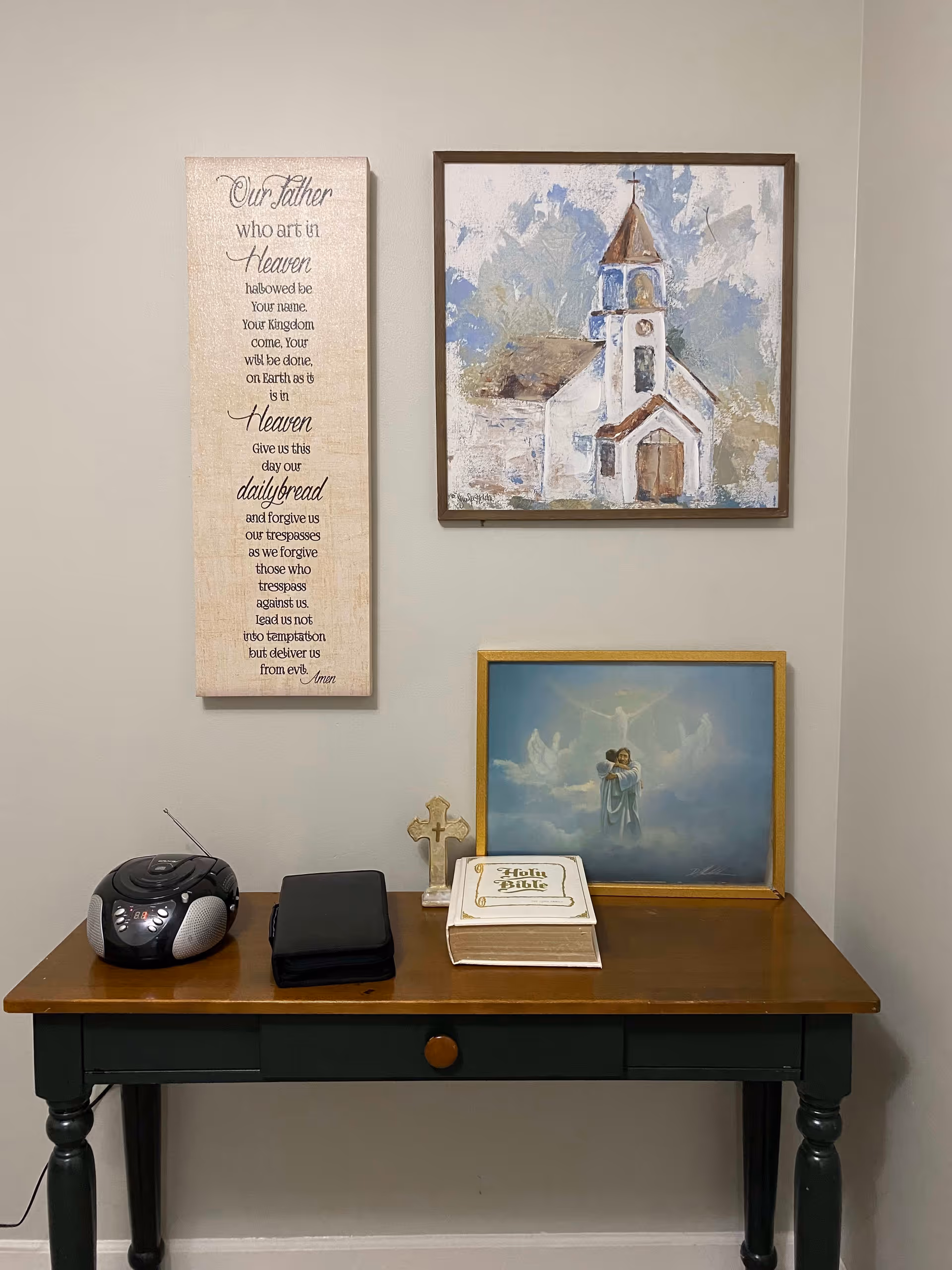 A wooden table against a beige wall with a small radio, a folded black item, a small cross, and a Holy Bible on top. Above the table are three framed items: a vertical plaque with the Lord's Prayer, a painting of a church, and a framed picture of an angel embracing a person.