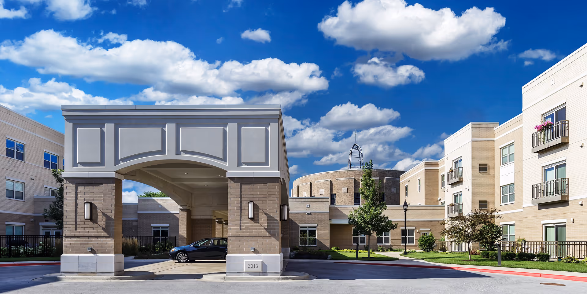 Exterior view of Mercy Circle Senior Living facility showing a covered entrance with brick pillars, a driveway with a parked car, and multi-story beige brick buildings with balconies under a blue sky with scattered clouds.