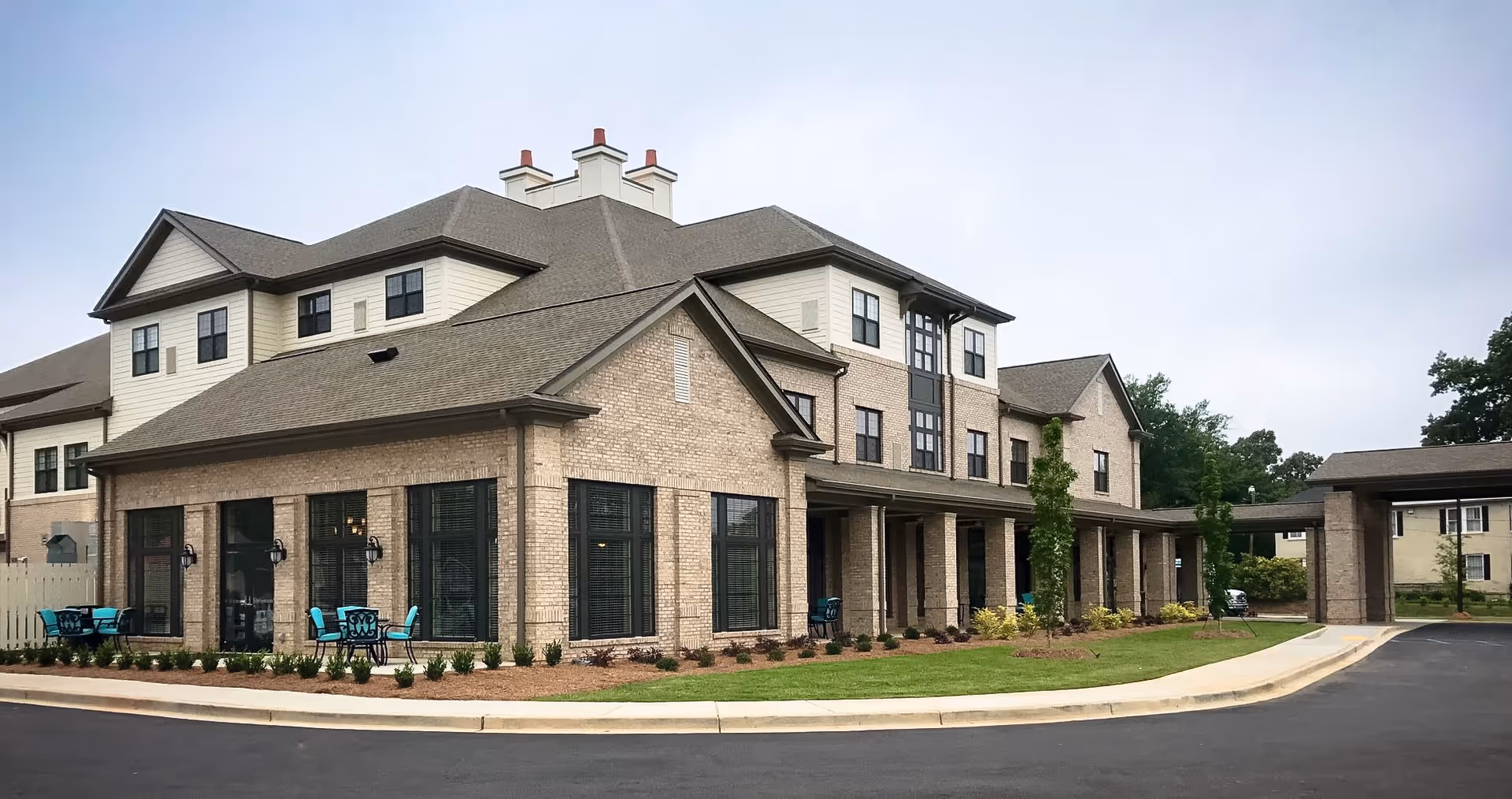 Exterior view of a multi-story senior living facility building with beige brick and light siding, large windows, and a covered entrance. There are small outdoor seating areas with turquoise chairs and tables, landscaped bushes, and a paved driveway in front.