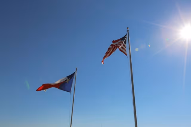 Two flagpoles against a clear blue sky with the sun shining brightly. One flagpole displays the Texas state flag and the other displays the United States flag.