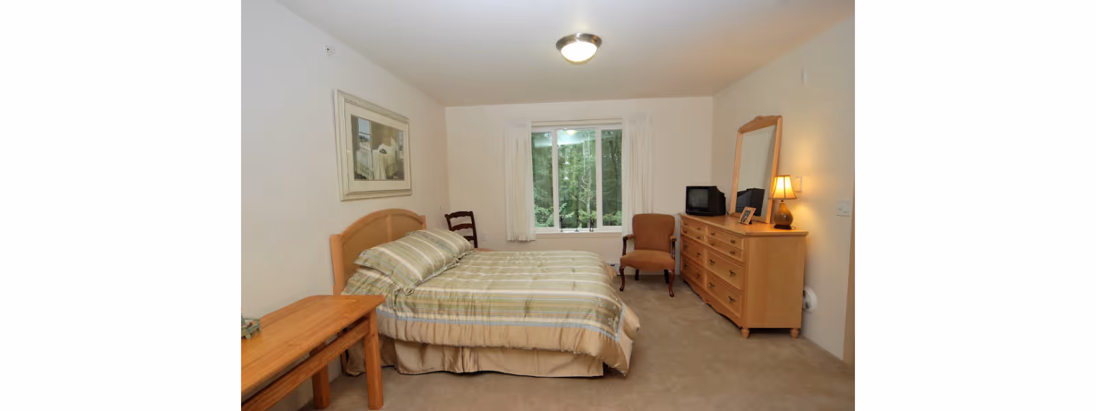 Light-filled bedroom with a bed, wooden dresser and mirror, armchair, and a window overlooking trees.