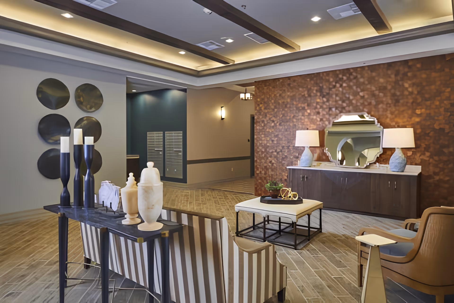 A modern living room area in an assisted living facility featuring a striped sofa, a cushioned ottoman with a tray holding decorative items, two armchairs, and a console table with two lamps and a decorative mirror above it. The room has a textured accent wall, recessed ceiling lights, and decorative wall art on a neutral wall.