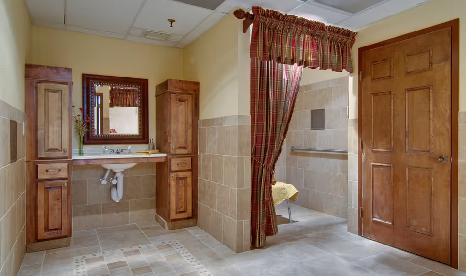 A bathroom area with beige tiled walls and floor, featuring a white sink with exposed plumbing, flanked by two wooden cabinets. A large mirror with a wooden frame hangs above the sink. To the right, a doorway with a wooden door and an adjacent open shower area with a plaid curtain and grab bars is visible.
