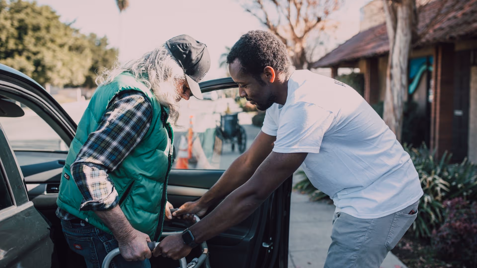 A man in a white t-shirt assisting an older man with long gray hair and a green vest as he gets out of a car using a walker, outdoors near a building with plants and trees in the background.
