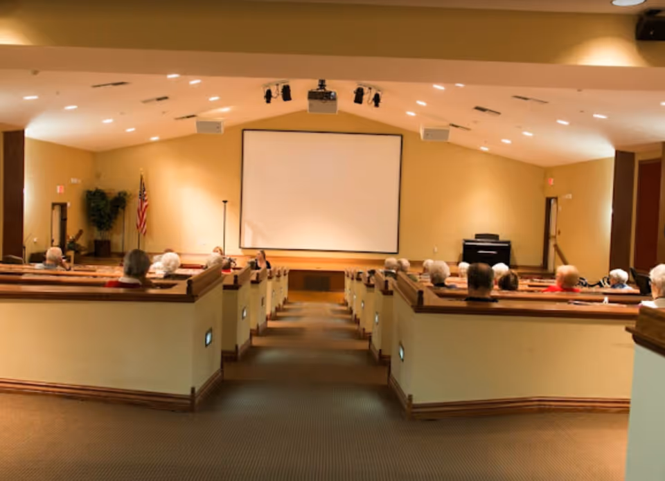A group of elderly people seated in a small auditorium or theater room facing a large blank projection screen. The room has beige walls, carpeted floor, and wooden pew-style seating with small lights along the aisle. An American flag and a potted plant are visible on the left side of the room.
