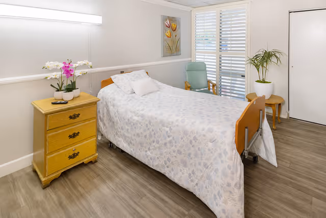 A single bed with a floral patterned bedspread in a senior living facility room. Next to the bed is a wooden nightstand with three drawers, holding a remote control and a small arrangement of white and purple orchids. A green armchair is positioned near a window with white plantation shutters. A potted plant sits on a small wooden table near a closed white door. The room has light-colored walls and wood-look flooring.
