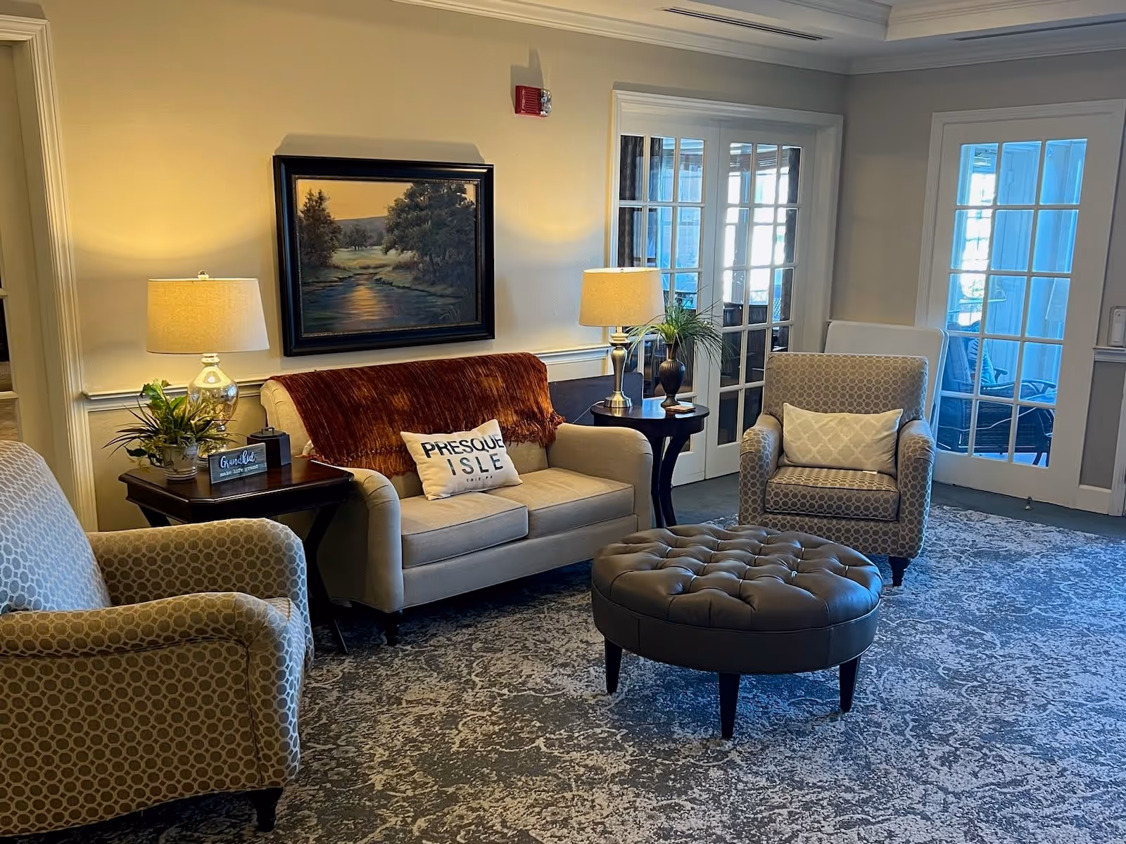 A cozy living room area with a beige loveseat adorned with a brown throw blanket and a pillow that reads 'PRESQUE ISLE'. There are two patterned armchairs, a round tufted ottoman in the center, two side tables with lamps and plants, and a framed landscape painting on the wall. French doors with glass panes lead to another room with seating visible through the glass.