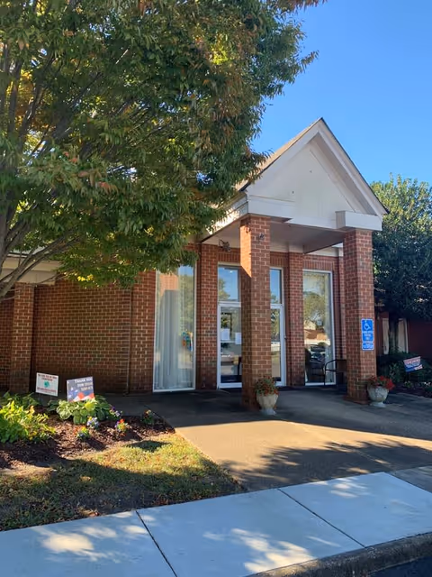 Entrance of a brick building with a covered porch supported by four columns. There are large windows and glass doors, a tree with green leaves on the left, flower beds, and a sidewalk leading to the entrance. A blue handicap parking sign is visible near the door.
