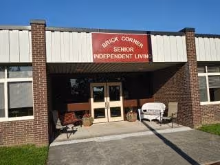 Front exterior view of Brick Corner Senior Independent Living facility showing a brick building with a covered entrance, a red sign above the door, two chairs and a bench on the porch, and a clear blue sky.