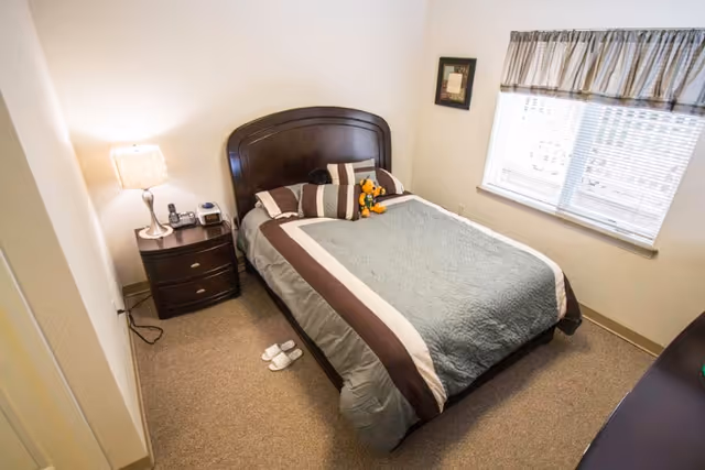 A small bedroom with a neatly made bed featuring a gray and brown comforter, a dark wooden headboard, and several pillows including a stuffed animal. There is a nightstand with a lamp, phone, and clock beside the bed, a window with blinds and a valance, and a pair of white slippers on the carpeted floor.
