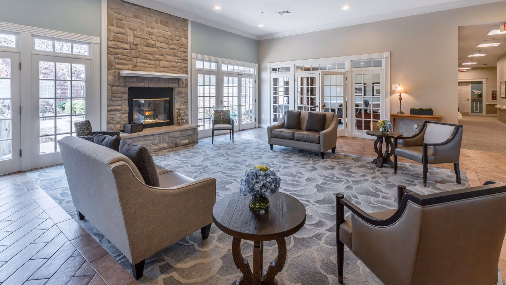 A bright and spacious living room area with a stone fireplace, multiple armchairs and sofas arranged around a round wooden coffee table with a flower vase. Large windows and glass doors allow natural light to fill the room, and a patterned area rug covers the floor.