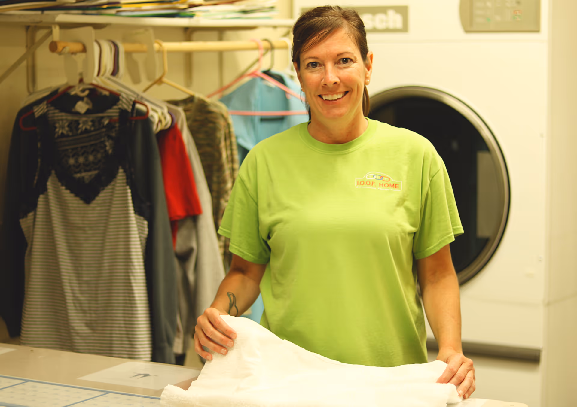 A woman wearing a green IOOF Home t-shirt is folding a white towel in a laundry room. Behind her, there are clothes hanging on a rack and a large front-loading dryer.