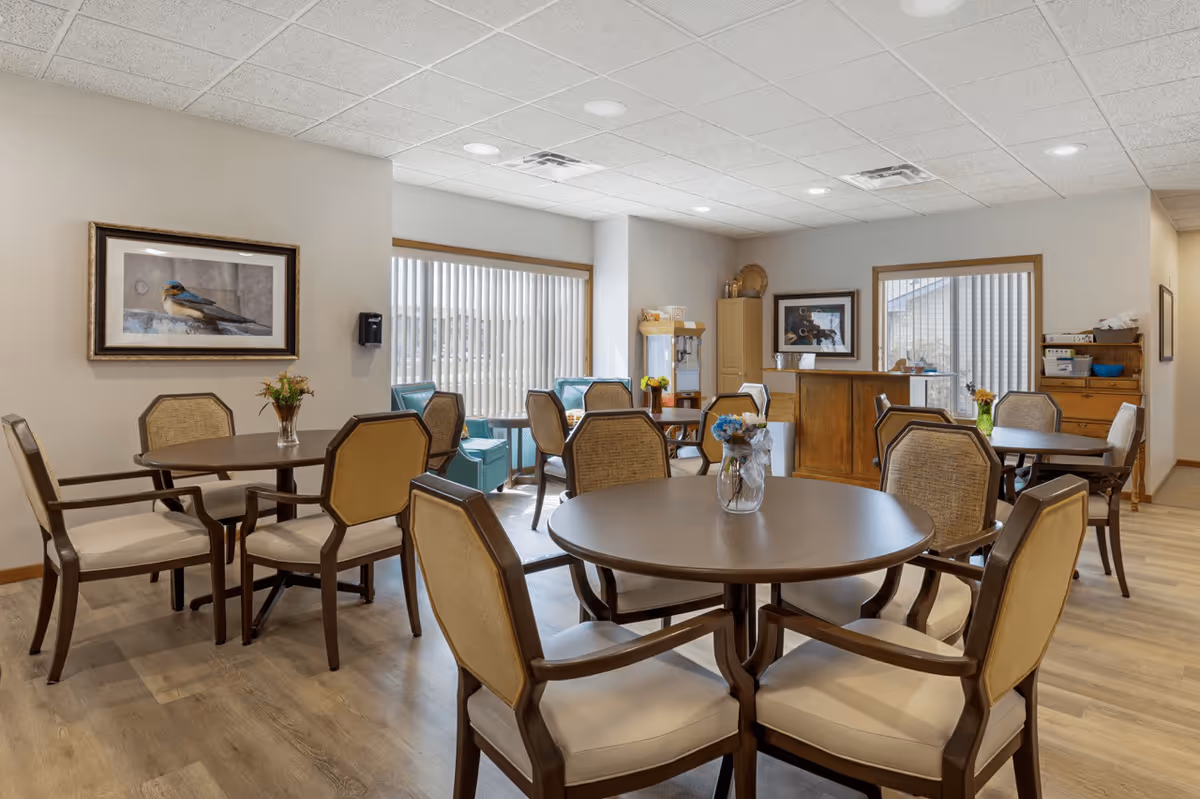 A bright dining room in a senior living facility with round wooden tables and cushioned chairs arranged neatly. The room has large windows with vertical blinds allowing natural light to enter. There are small flower vases on the tables, framed artwork on the walls, and a wooden cabinet and popcorn machine in the background.