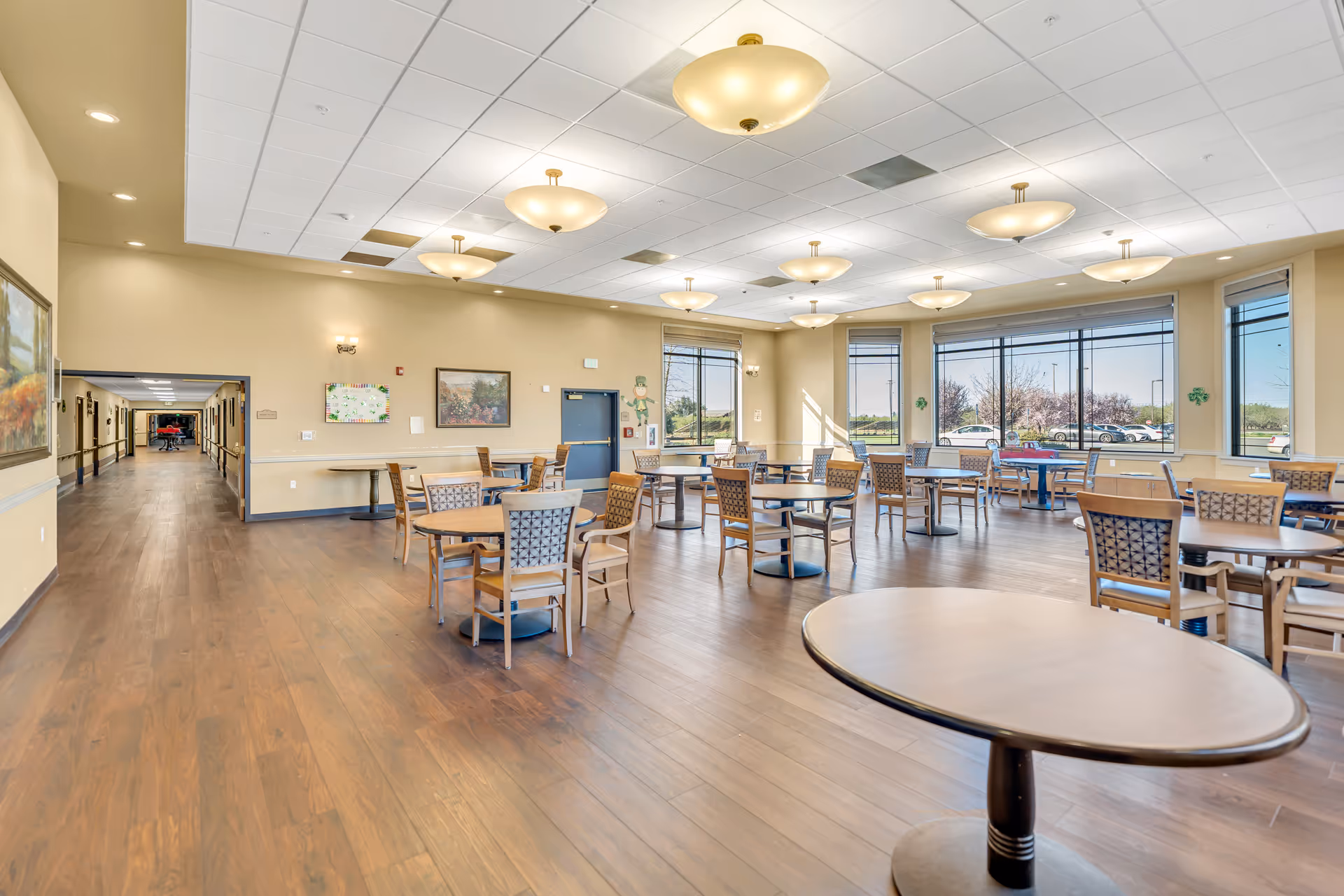 A spacious dining room with multiple round tables and chairs arranged neatly on a wooden floor. Large windows on one side allow natural light to fill the room, and several ceiling lights provide additional illumination. The walls are decorated with paintings and a bulletin board. A hallway extends from the dining area, and outside the windows, parked cars and trees are visible.