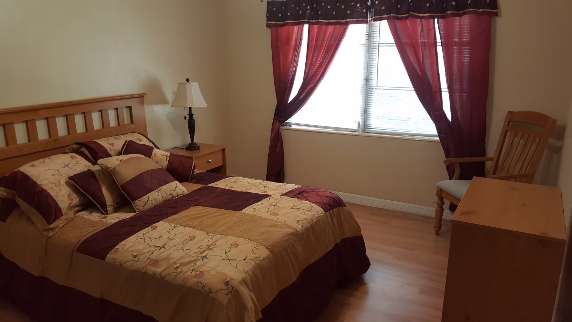 A bedroom with a wooden bed frame and a bed made with a beige and burgundy floral patterned comforter and matching pillows. There is a wooden nightstand with a lamp on it next to the bed. A window with burgundy curtains and a valance lets in natural light. A wooden chair and a wooden dresser are also visible in the room with light-colored walls and wood flooring.