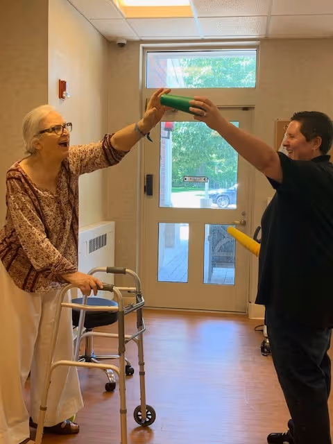 An elderly woman using a walker is smiling and reaching out to touch a green cylindrical object held by a younger person in a hallway with a glass door in the background.