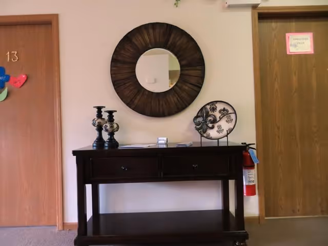 A hallway area with a dark wooden console table against a light-colored wall. On the table are two decorative candle holders, a decorative plate on a stand, and some papers. Above the table is a round mirror with a dark wooden frame. To the left is a door labeled with the number 13 and colorful paper hearts attached. To the right is another door with a pink sign and a fire extinguisher mounted on the wall beside it.