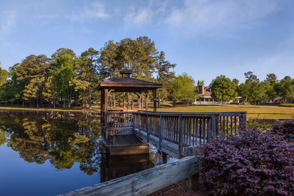 A wooden gazebo on a pier extending over a calm pond surrounded by trees and greenery, with a clear blue sky overhead and a building visible in the background.