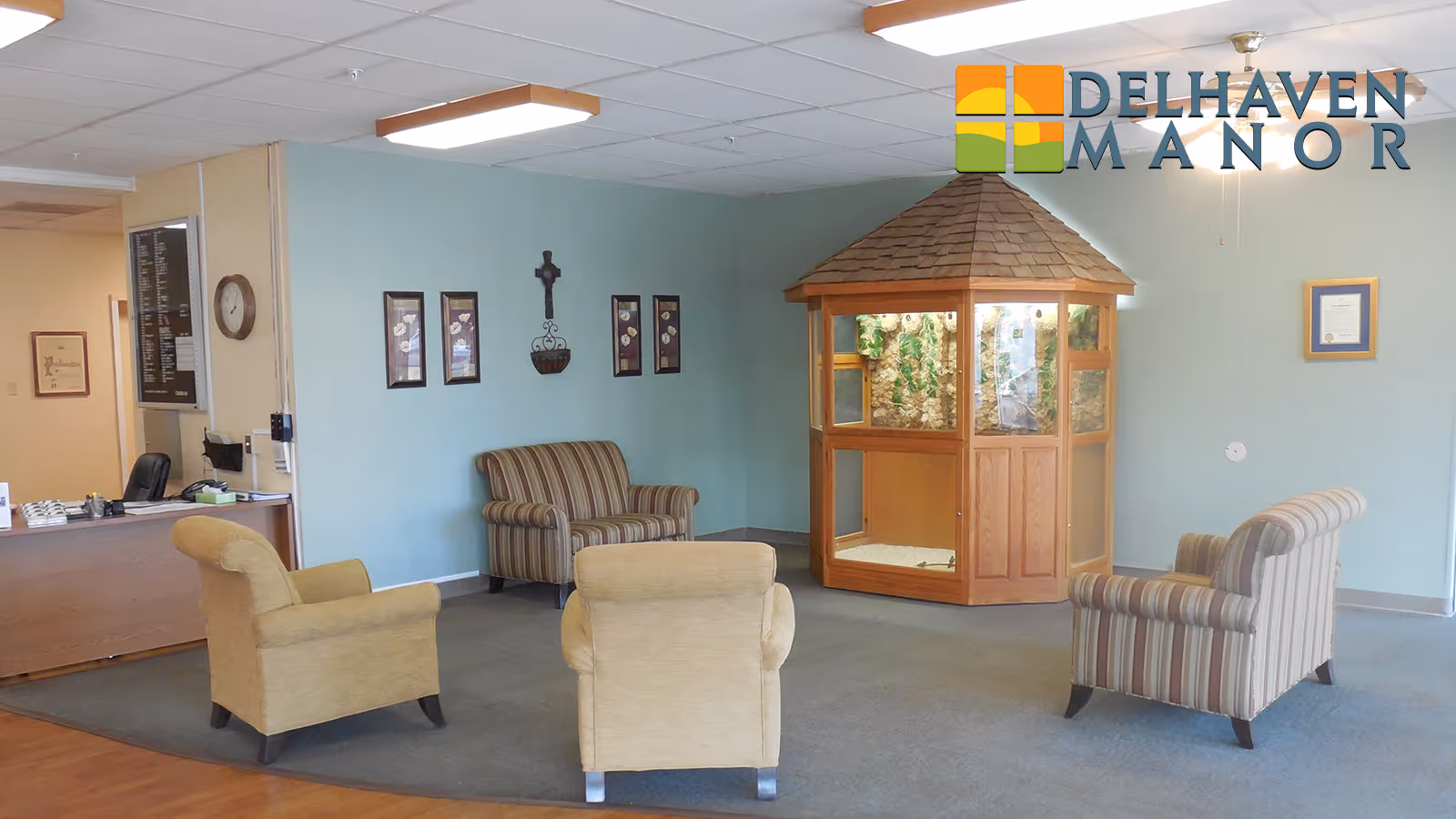 A nursing center lobby with four upholstered armchairs arranged around a wooden display case with a shingled roof. The walls are light blue with framed pictures and a cross hanging. A reception desk is visible to the left. The Delhaven Manor logo is in the top right corner.
