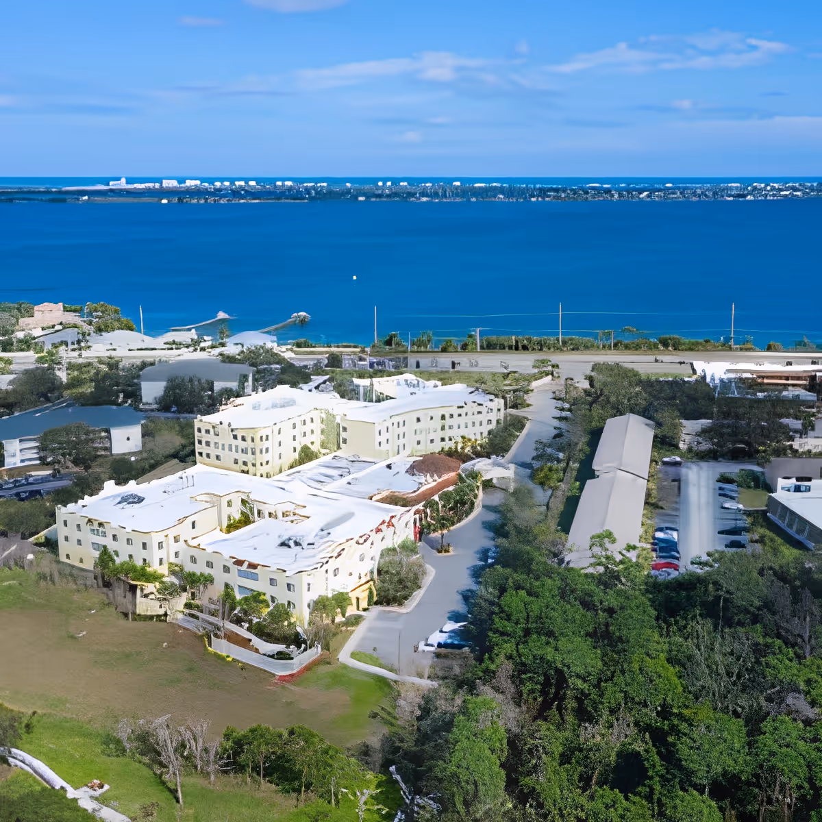 Aerial view of Discovery Village At Melbourne senior living facility near a large body of water, showing multiple buildings, parking areas, and surrounding greenery under a blue sky.