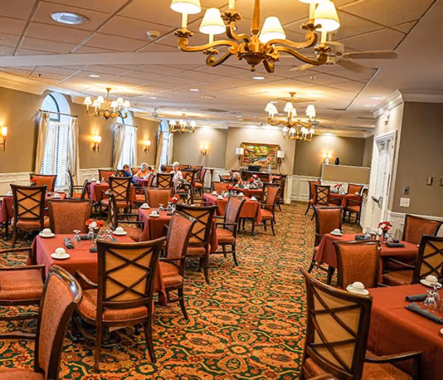 A spacious dining room with multiple tables covered in red tablecloths, each set with white cups and napkins. The room features patterned carpet, wooden chairs with cushioned seats, chandeliers hanging from the ceiling, and wall sconces providing warm lighting. Several elderly people are seated at tables near the windows on the left side of the room.