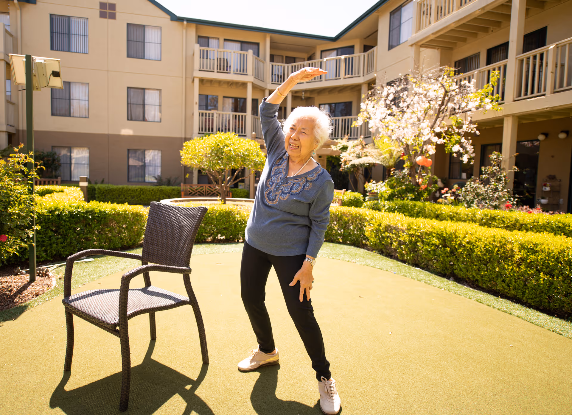 An elderly woman is standing outdoors in a courtyard of a senior living facility, stretching with one arm raised above her head and the other resting on her thigh. She is smiling and appears to be exercising next to a wicker chair. The courtyard is surrounded by a beige multi-story building with balconies, green hedges, trees, and flowering plants under a sunny sky.