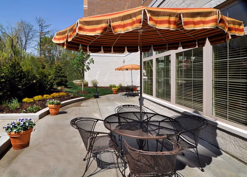 Outdoor patio area with metal table and chairs under a large striped umbrella. There are potted flowers and landscaped greenery along the concrete walkway next to a building with large windows and blinds.