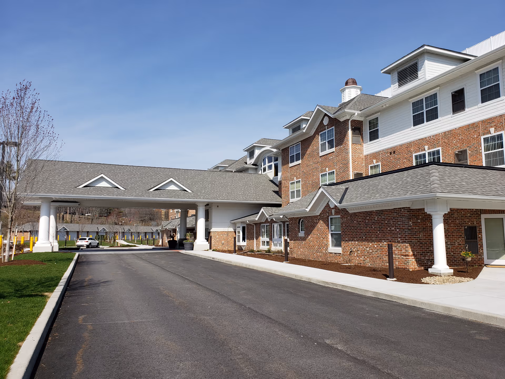Driveway leading to the covered entrance of a multi-story brick and white-siding retirement community building under a clear blue sky.