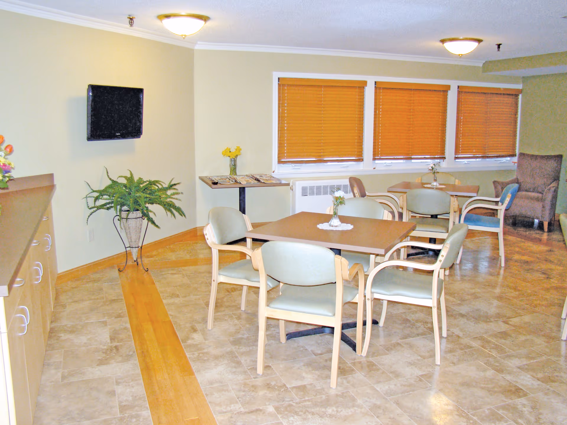 A well-lit common area with several tables and chairs arranged for seating. The room features beige tiled flooring with a wooden strip, light-colored walls, three windows with closed wooden blinds, a wall-mounted TV, a potted plant, and a small table with magazines and a vase with yellow flowers. There is also a comfortable armchair in the corner.