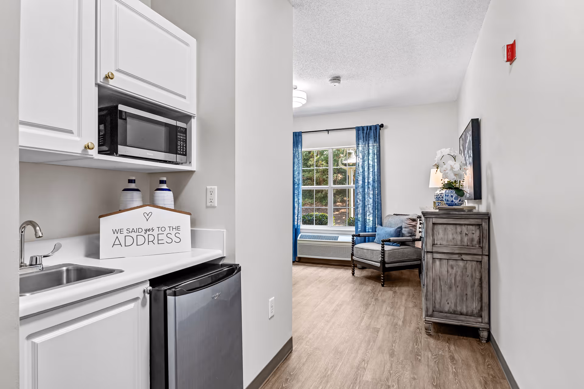 A small kitchenette area with a sink, microwave, and mini refrigerator on the left side. A decorative sign on the counter reads 'We said yes to the address.' Beyond the kitchenette, there is a cozy sitting area with a cushioned chair, a wooden cabinet with a lamp and flowers, and a large window with blue curtains letting in natural light.