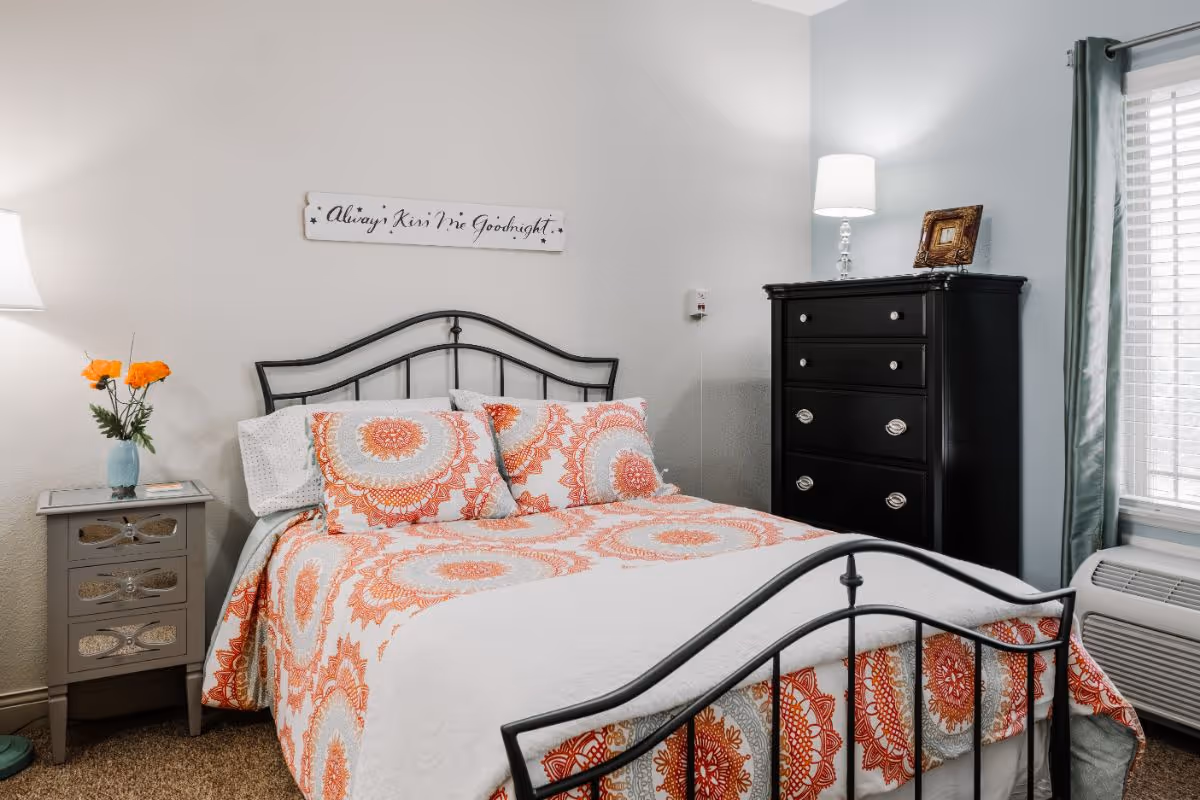 A cozy bedroom with a black metal bed frame, a bed dressed in white and orange patterned bedding, and matching pillows. To the left of the bed is a small gray nightstand with a vase holding orange flowers and a lamp. On the right side, there is a tall black dresser with a lamp and a small framed picture on top. A window with gray curtains and blinds is visible on the right wall. Above the bed, a decorative sign reads 'Always Kiss Me Goodnight.'