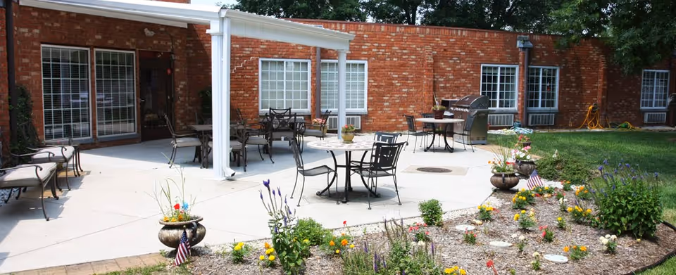 Outdoor patio area with several metal tables and chairs on a concrete surface, adjacent to a red brick building with multiple windows. There are flower beds with colorful flowers and small American flags in the foreground, and a barbecue grill near the building. Trees and greenery surround the area.