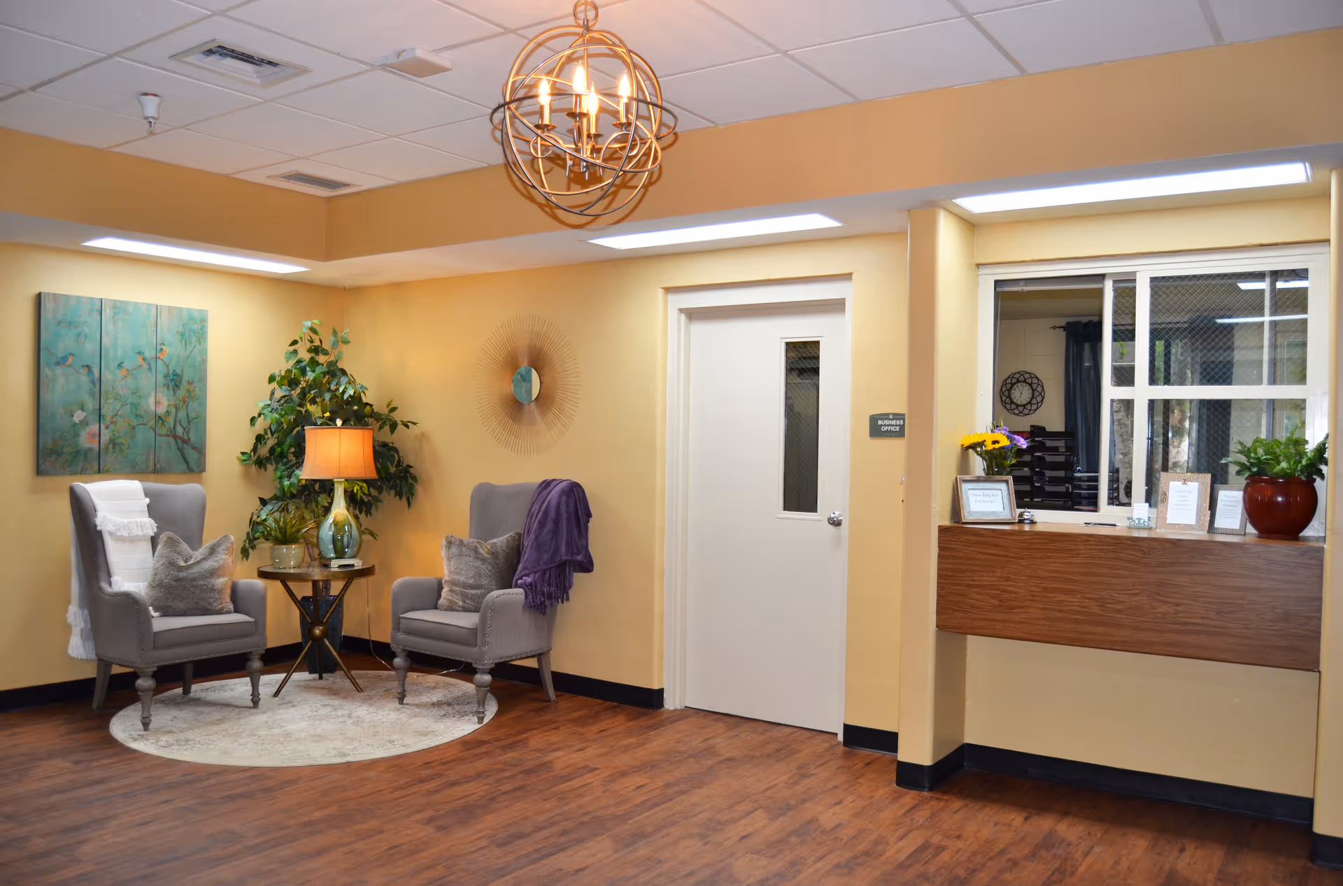 A cozy seating area in a senior living facility with two gray armchairs, a small round table with a lamp and plant, wall art, and a decorative mirror. To the right is a reception window with a wooden counter and a door labeled 'Business Office'. The walls are painted a warm yellow and the floor has wood-like flooring.