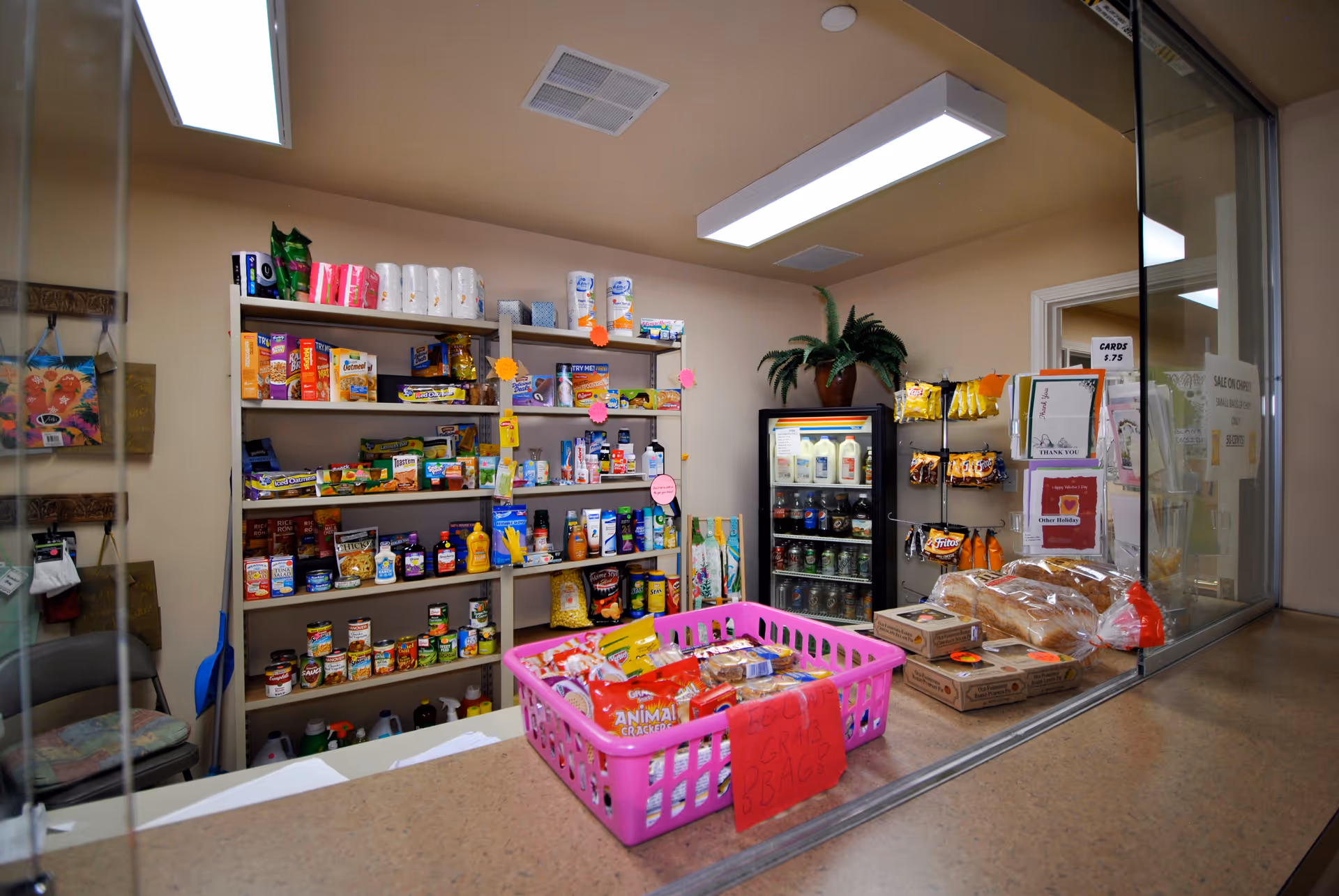 Interior view of a small convenience store or snack shop area within a facility, featuring shelves stocked with various food items, snacks, and household products. A pink basket filled with snacks is on the counter, along with loaves of bread and boxes. A refrigerated display case with beverages is visible in the background, and a glass partition separates the counter from the customer area.