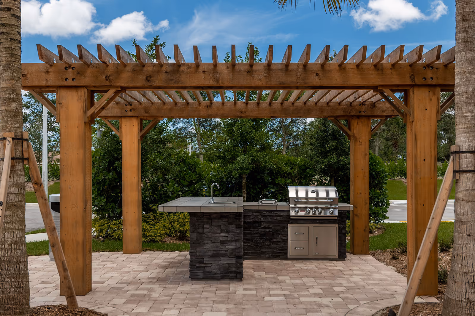 Outdoor grilling area with a stone and tile countertop, stainless steel grill, and sink under a wooden pergola surrounded by greenery and palm trees.
