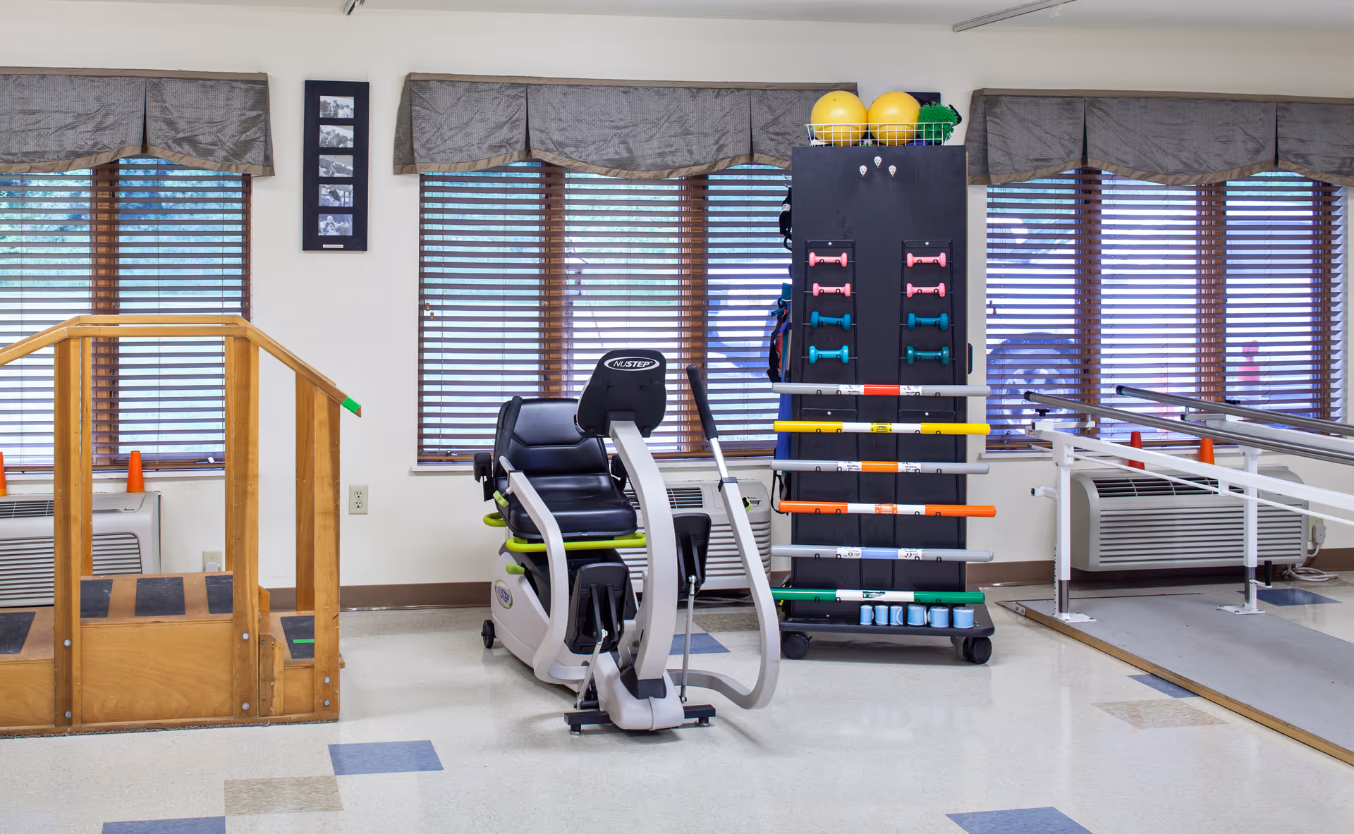 A physical therapy or rehabilitation room with exercise equipment including a recumbent exercise bike, a wooden staircase with handrails for walking practice, parallel bars for gait training, and a rack holding colorful dumbbells, exercise balls, and other fitness accessories. The room has large windows with blinds and valances.