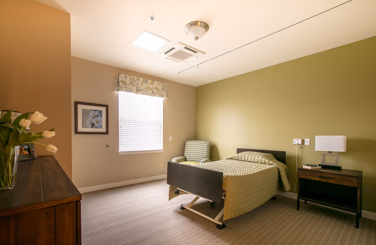 A simple, well-lit bedroom with a single bed covered in a light green quilt. There is a striped armchair in the corner next to the bed, a wooden nightstand with a modern lamp, and a wooden dresser with a vase of white tulips and a framed photo. The walls are painted in beige and light green, and a window with blinds and a floral valance lets in natural light.