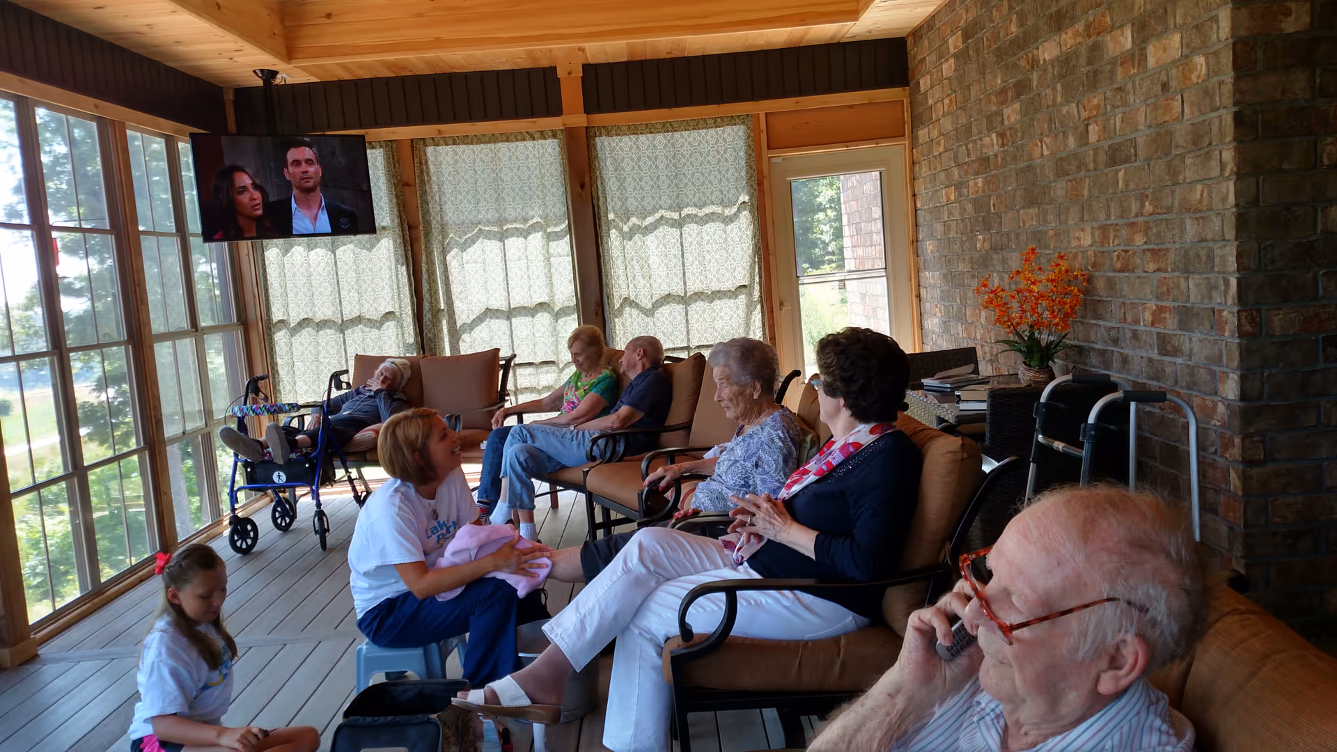 A group of elderly people and a caregiver are seated in a sunroom with large windows and wooden ceiling. Some are sitting on cushioned chairs while one elderly man is using a walker. The caregiver is interacting with a young girl sitting on the floor. A TV is mounted on the wall showing a scene from a show or movie. The room has a cozy atmosphere with brick walls and a vase of orange flowers on a side table.