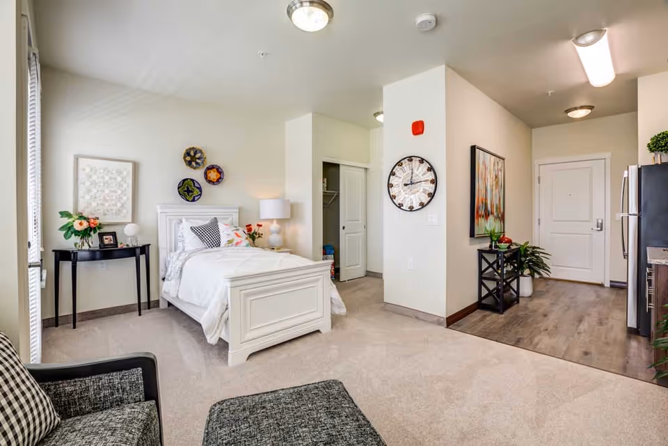 A cozy bedroom interior featuring a white bed with decorative pillows, a bedside table with a lamp, and a small table with plants. The room has light-colored walls and a modern clock on the wall, with a view of the entrance door.