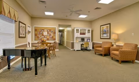 A spacious senior living common area with a black grand piano on the left, a round wooden table with chairs, a bulletin board on the wall, a white desk with a chair, two upholstered armchairs with a side table and lamp, and framed artwork on the walls. The room has beige walls and carpeted floor with ceiling lights and a ceiling fan.