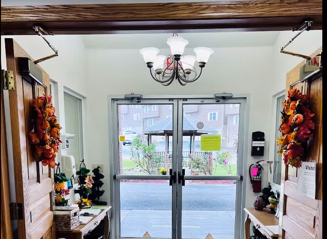View through double glass doors leading outside from an interior hallway decorated with autumn wreaths on wooden doors and small tables with flowers and decorative items on each side.