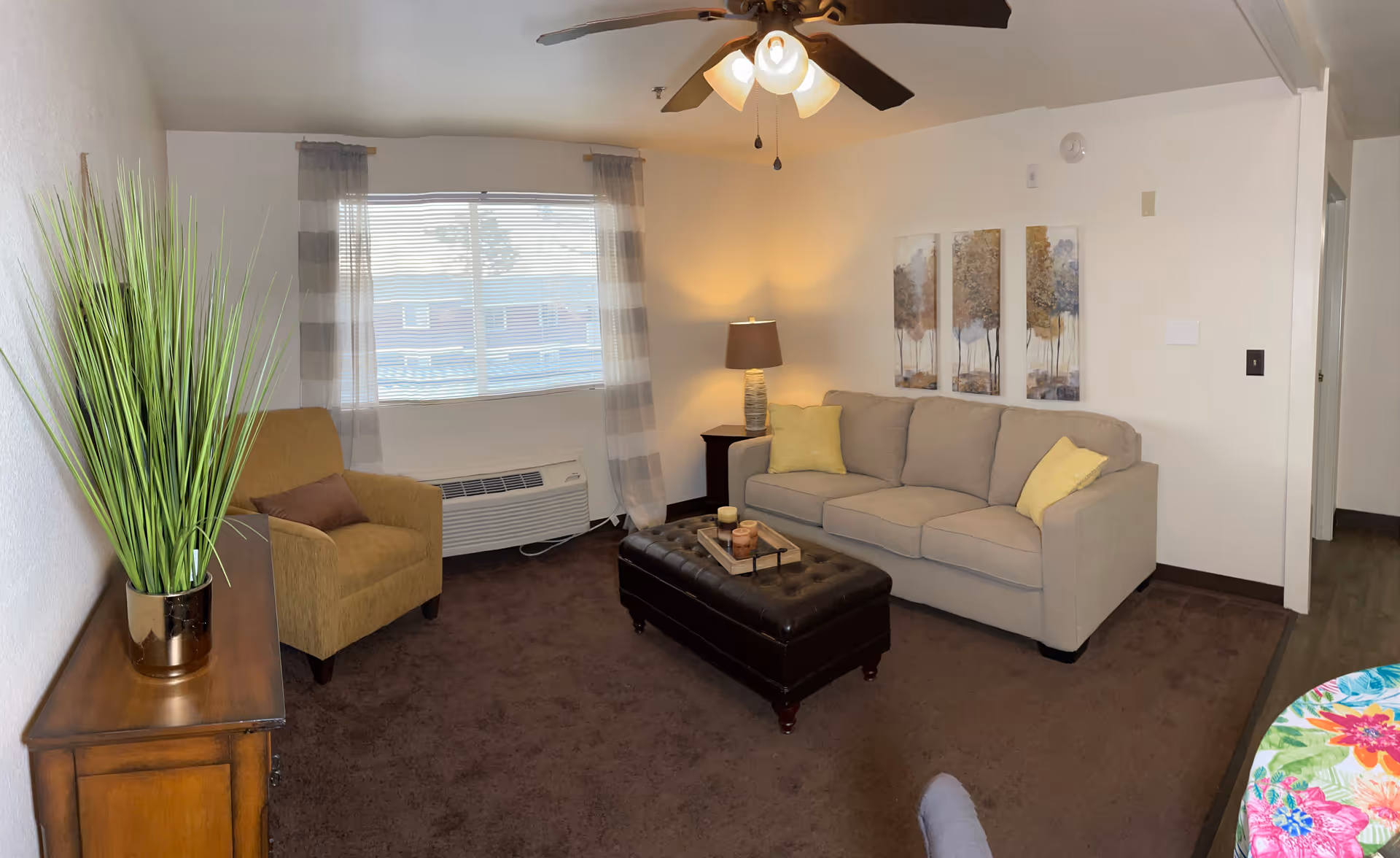 A cozy living room with a beige sofa adorned with yellow pillows, a brown armchair, a dark brown tufted ottoman serving as a coffee table with candles on a tray, a wooden side table with a lamp, a large window with striped curtains, a ceiling fan with lights, and a wooden cabinet with a tall green plant in a gold pot.