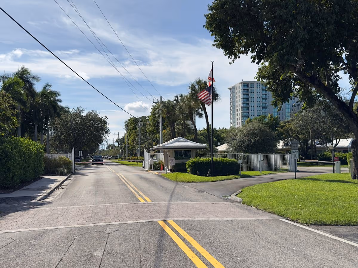 Street leading to a gated entrance with a guardhouse, flagpoles, palm trees and a high-rise building in the background.