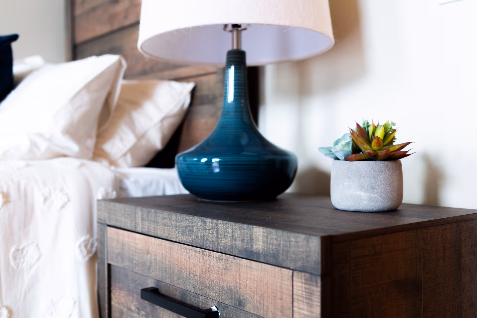 Close-up of a wooden bedside table with a blue ceramic lamp and a small potted succulent plant, next to a bed with white bedding and pillows.